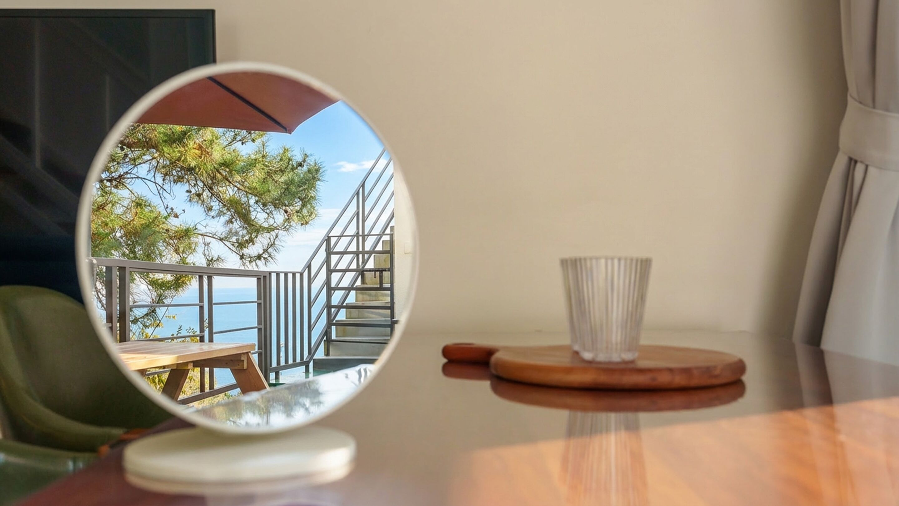 A round mirror reflecting a balcony with a wooden table and a glass cup on a wooden coaster.