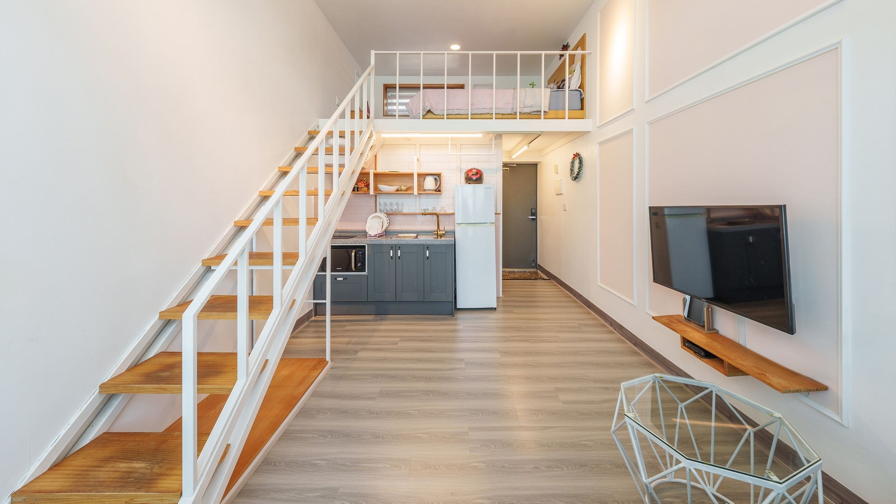 A modern apartment with a wooden staircase, a kitchen with a white refrigerator, and a flat-screen TV mounted on the wall.