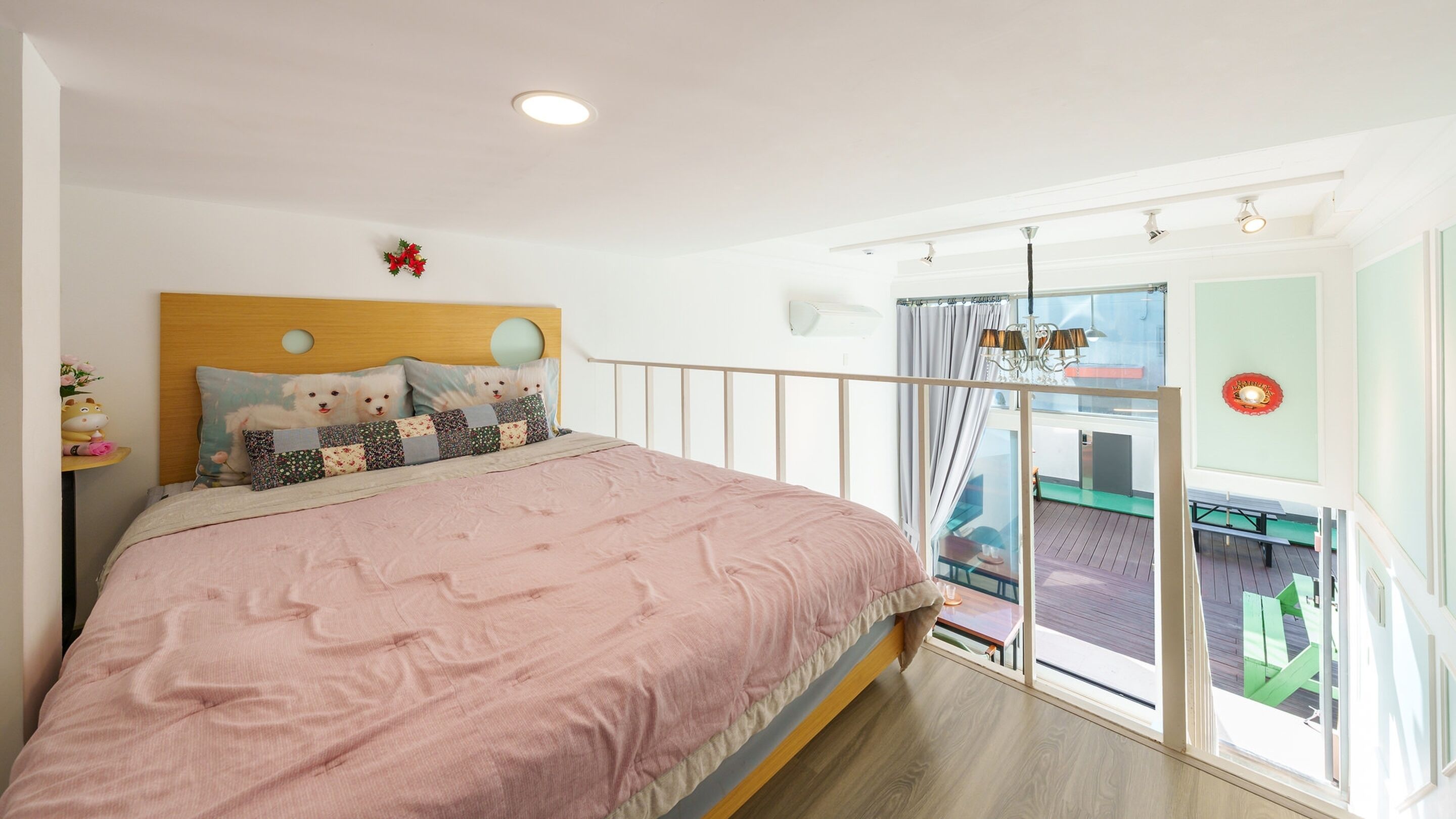A bedroom with a bed, a nightstand, a window with curtains, a view of the outside balcony, and a wall-mounted air conditioning unit.