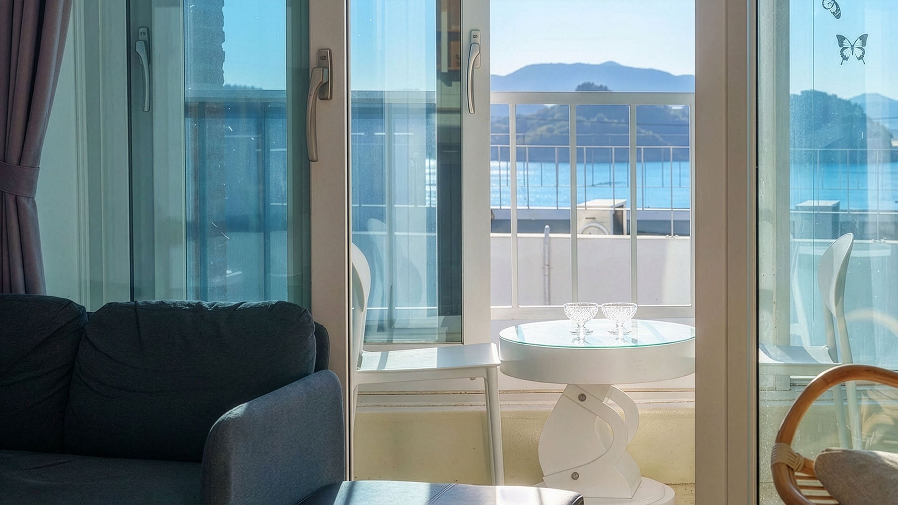 A balcony with a glass table and chairs, a sofa, and a view of the sea and mountains.