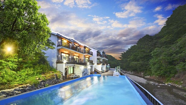A multi-story hotel with a swimming pool, located beside a river, surrounded by greenery. - Gyeongju Green Valley Spa Pension (Gyeongju)