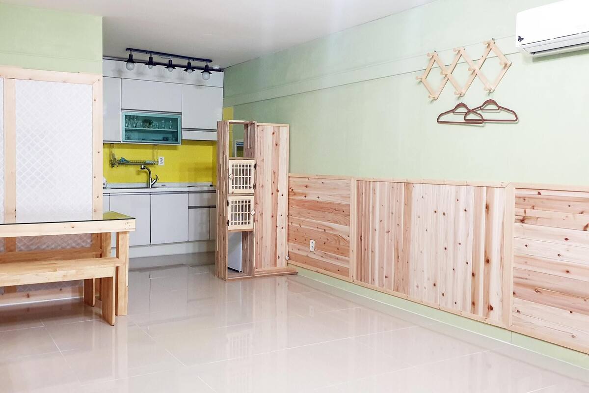 A room with a wooden bench, a small table, a kitchen area with a sink and cabinets, and a wall-mounted air conditioner.