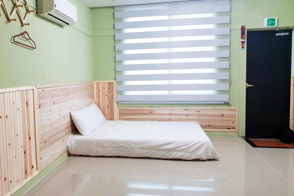 A room with a wooden headboard, a bed with white bedding, a window with blinds, and a door with an emergency exit sign.