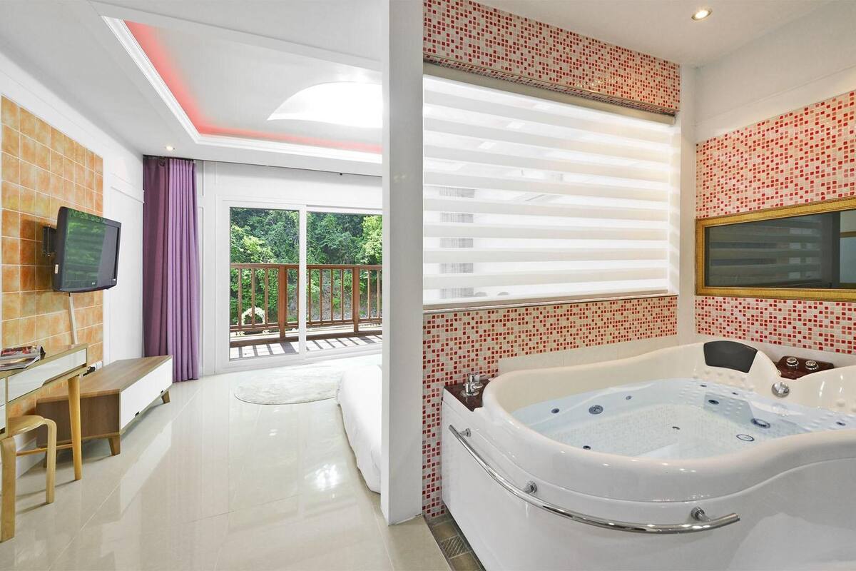 A modern bathroom with a large bathtub, a television, and a view of greenery outside.