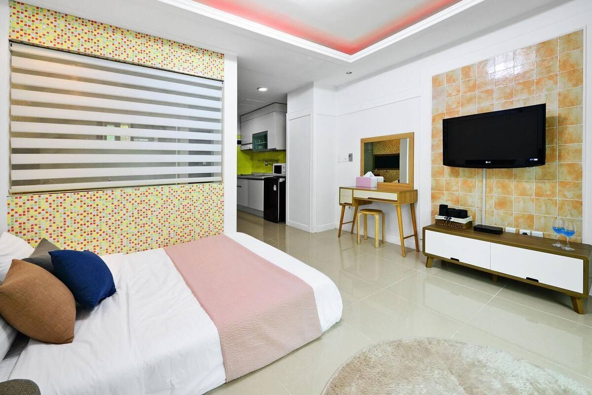 A modern hotel room with a bed, a desk, a TV, and a kitchen area in the background.