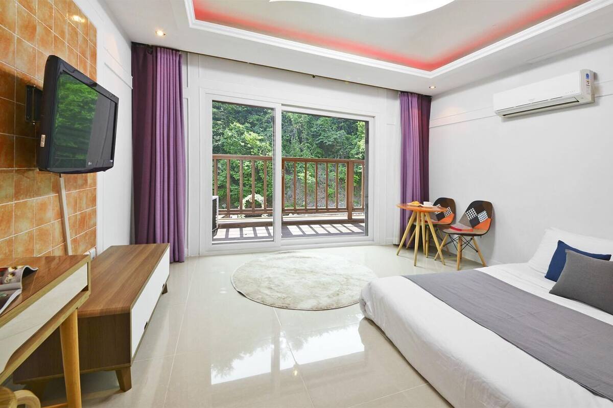 A modern hotel room with a large bed, a desk, a TV, and a balcony with a view of greenery.