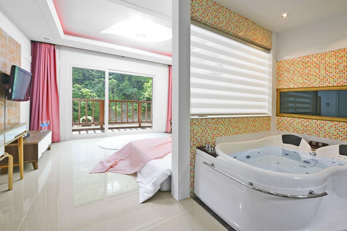 A spacious bedroom with a large window, a balcony, a jacuzzi, and a TV.