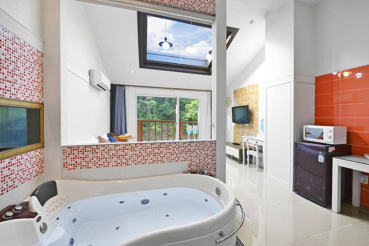 A modern bathroom with a large bathtub, a skylight, a TV, and a kitchen area with orange tiles.