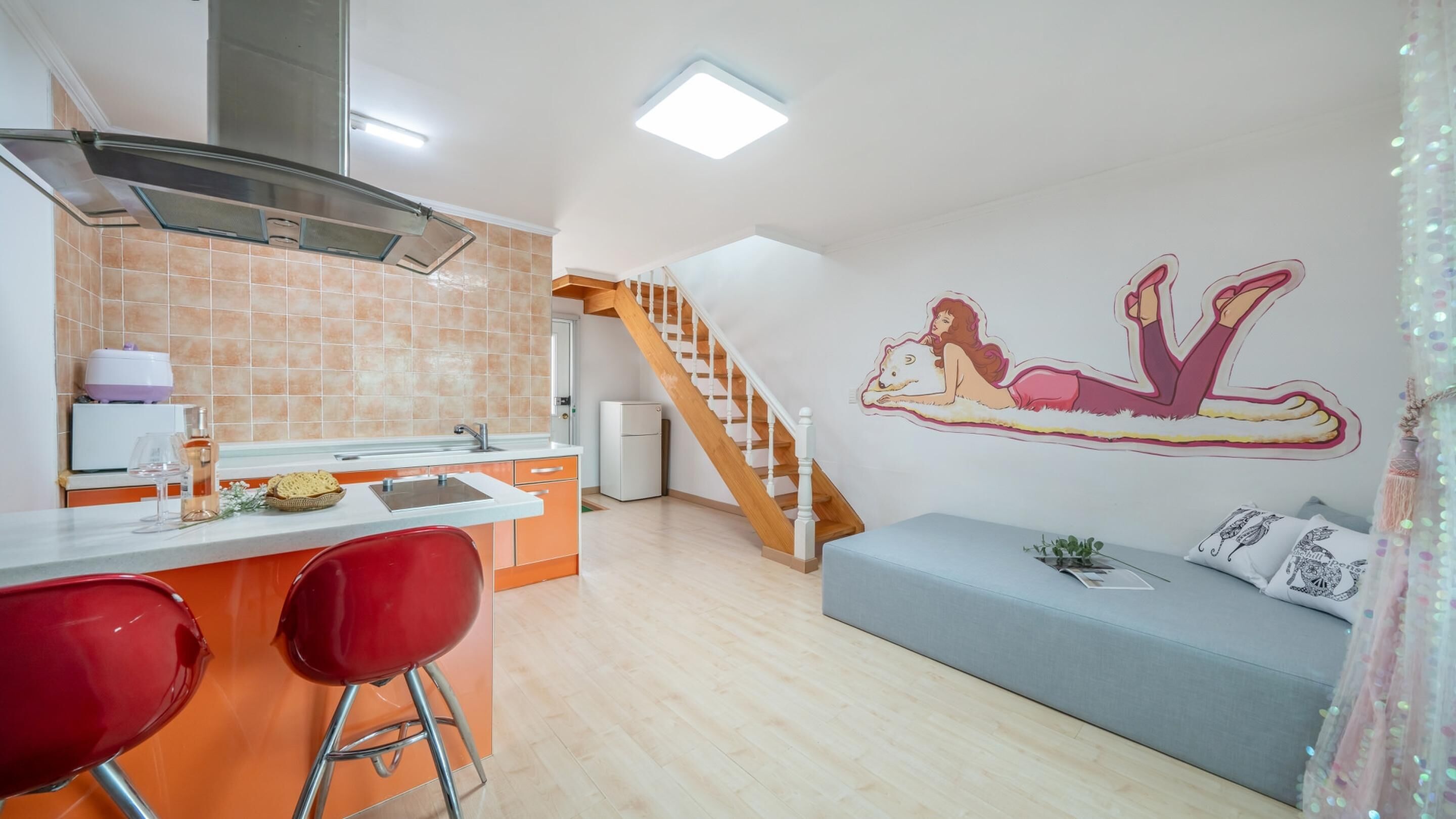 A modern kitchen with a central island, a wooden staircase, and a living area with a sofa and decorative pillows.