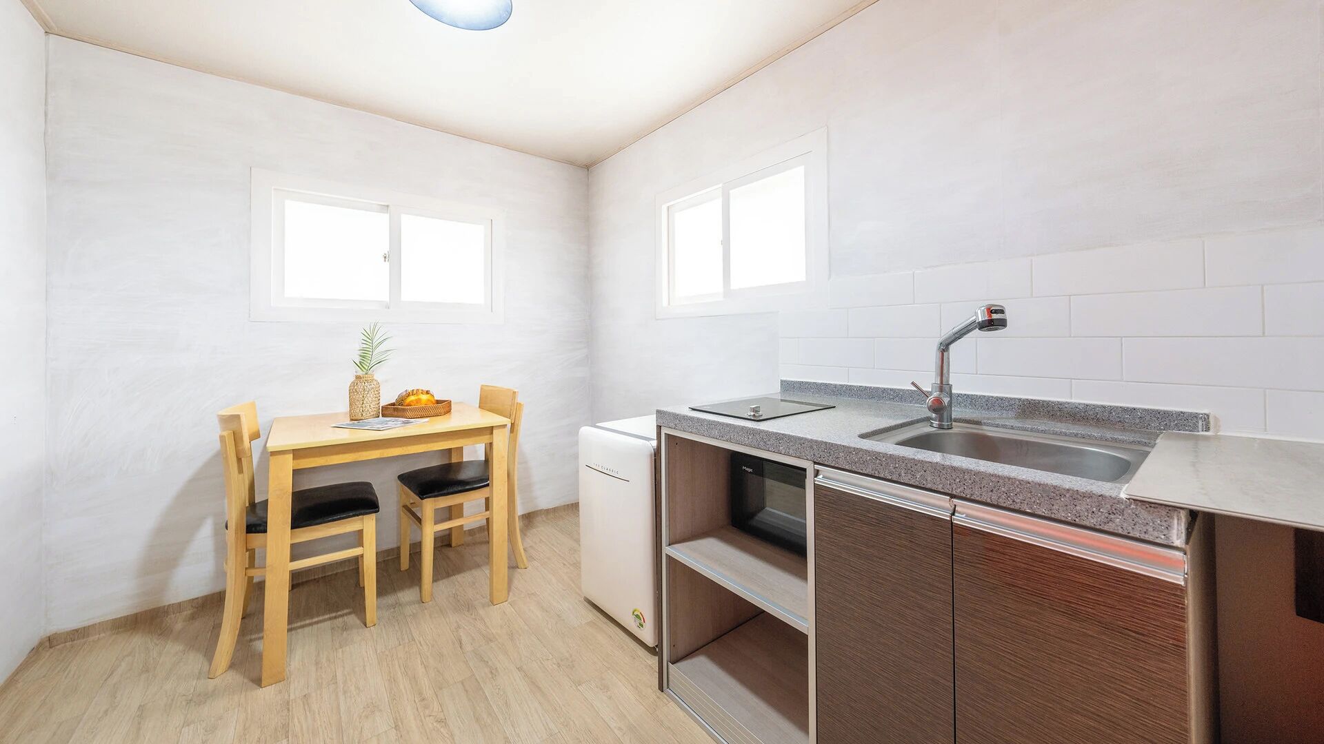 A compact kitchen with a wooden table, two chairs, a built-in dishwasher, and a sink with a modern faucet.