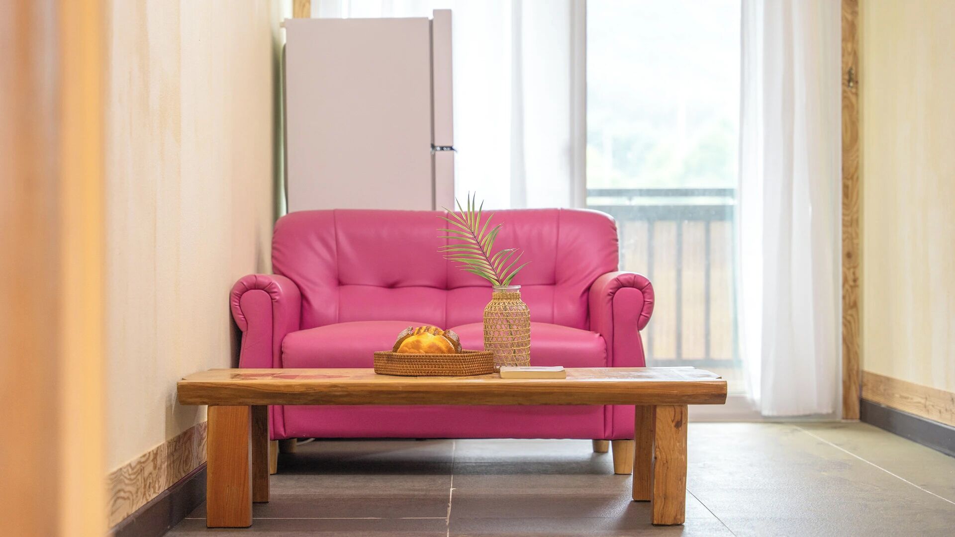 A pink sofa, a wooden table, and a vase with a plant in a room with a window.