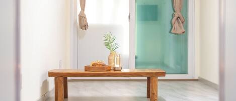 Un banc en bois, un panier tressé et un vase en verre contenant une plante, le tout dans une pièce dont le sol est recouvert de carreaux et qui possède une porte en verre.