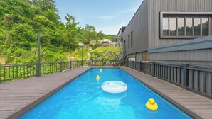 Una piscina con cubierta de madera, rodeada de vegetación y un edificio moderno.
