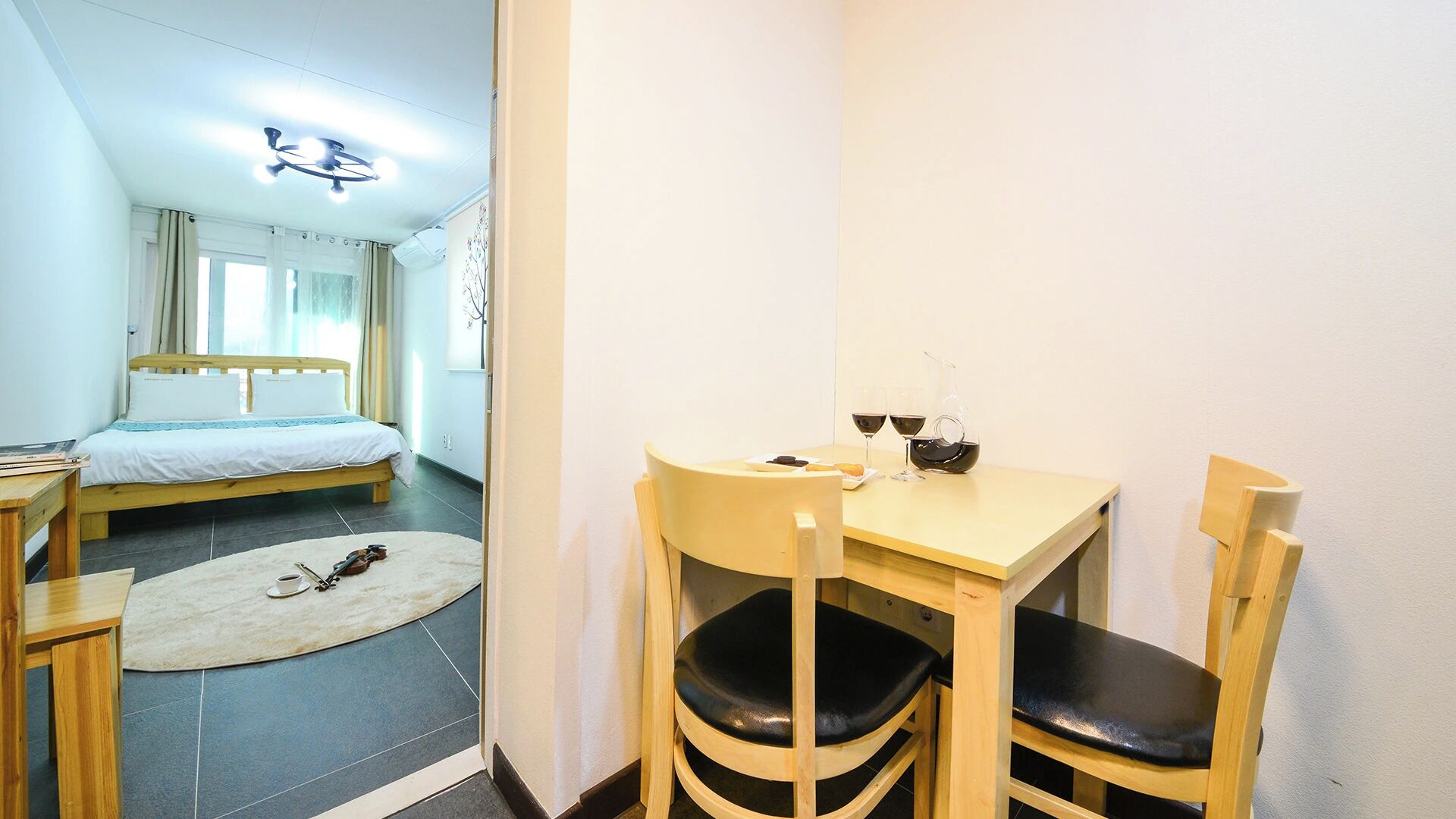 A hotel room with a bed, a desk with chairs, a small rug, and a violin.
