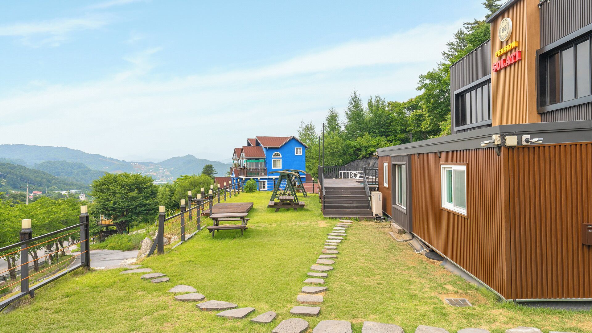 A residential area with a blue house, a wooden building with a sign, and a stone pathway leading to a wooden deck.