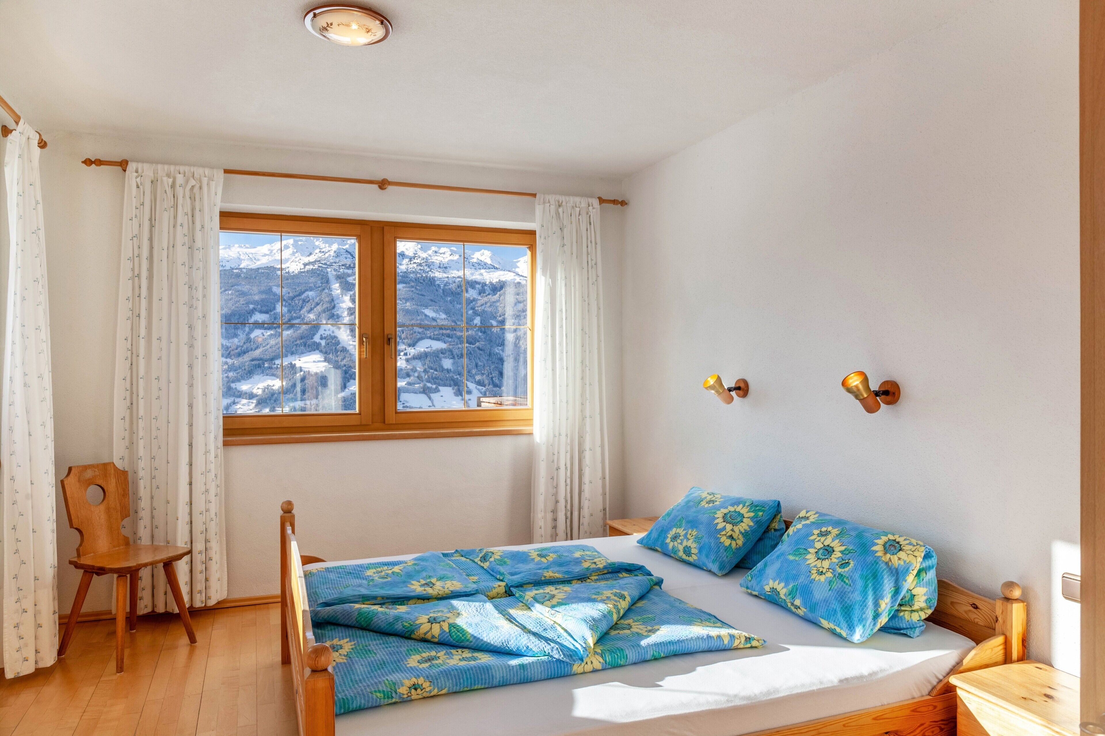 A bedroom with a bed, wooden chair, window with curtains, and a view of snow-covered mountains.