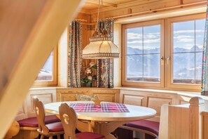 Un comedor acogedor con una mesa redonda, sillas de madera y un banco, con vistas a la montaña a través de amplias ventanas.