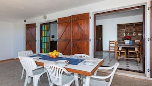 Un comedor con una mesa preparada para cuatro personas, sillas de plástico blancas, un cuenco de fruta y un mantel individual azul.