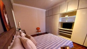 Un dormitorio moderno con cama, mesita de noche, sillón y almacenamiento integrado con televisor.