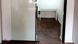 Una habitación con puerta corrediza de vidrio que da a un baño con inodoro blanco y un lavamanos pequeño. El piso es de madera y las paredes son blancas.