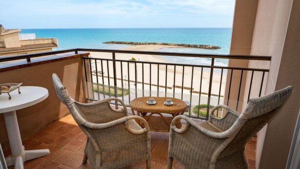 A balcony with wicker chairs and a small round table, overlooking a beach with a rocky breakwater.