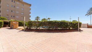 A building with a flat roof and multiple windows, surrounded by a paved area with a low hedge and palm trees.