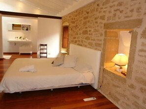 Un dormitorio con una cama grande, una pared de piedra, suelo de madera, una mesita de noche con lámpara, y un baño con lavamanos y espejo.