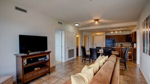 A spacious living room with a large flat-screen TV, a wooden entertainment unit, a brown sofa with patterned cushions, and a kitchen area in the background. - Mountainview getaway at Playa Del Sol (Kelowna)