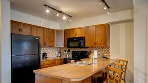 A kitchen with wooden cabinets, a black refrigerator, a microwave, a stove, and a sink. There are two wooden chairs with striped cushions. - Mountainview getaway at Playa Del Sol (Kelowna)