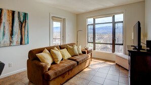 A living room with a brown sofa, a painting on the wall, and a large window with a view of mountains. - Mountainview getaway at Playa Del Sol (Kelowna)