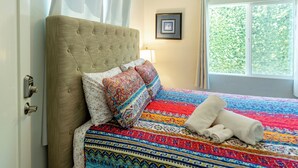 Un dormitorio con cabecera capitonada, ropa de cama estampada, una ventana con vista a follaje y un cuadro en la pared.