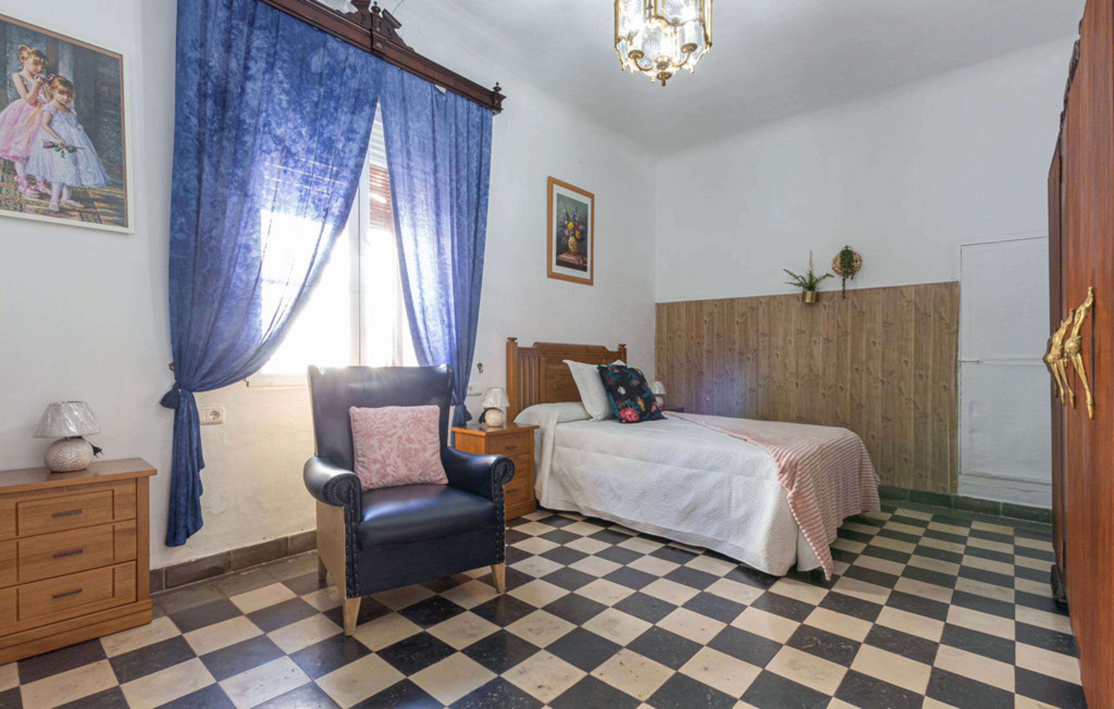 Una camera da letto con pavimento a scacchi, un letto con cuscino floreale, un comodino, una cassettiera e un quadro appeso al muro.
