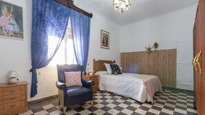 Een slaapkamer met een zwart-wit betegelde vloer, een bed met een bloemenkussen, een nachtkastje, een dressoir en een schilderij aan de muur.