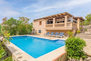 Vue sur une maison à deux étages avec balcon, une piscine et des chaises longues, le tout vu depuis le bord de la piscine.