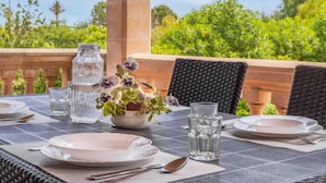 Una mesa al aire libre con platos, cubiertos, vasos y un florero.