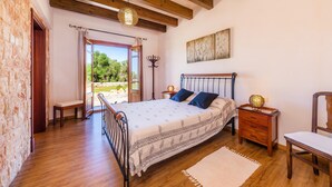 Ein Schlafzimmer mit einem hölzernen Bett, einer Bank, einem Nachttisch, einem Sessel und Blick auf Grünflächen.