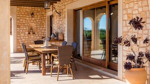 Eine Terrasse mit Steinwänden, einem Holztisch und Korbstühlen, einem Blumentopf und einer Glastür, die in einen Außenbereich führt.