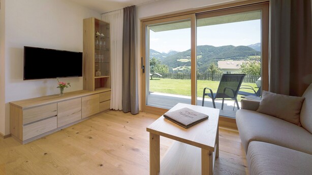 Un salon doté d’un téléviseur à écran plat, de meubles en bois, d’une table avec un livre et d’une porte-fenêtre coulissante donnant sur un espace extérieur avec vue sur les collines.