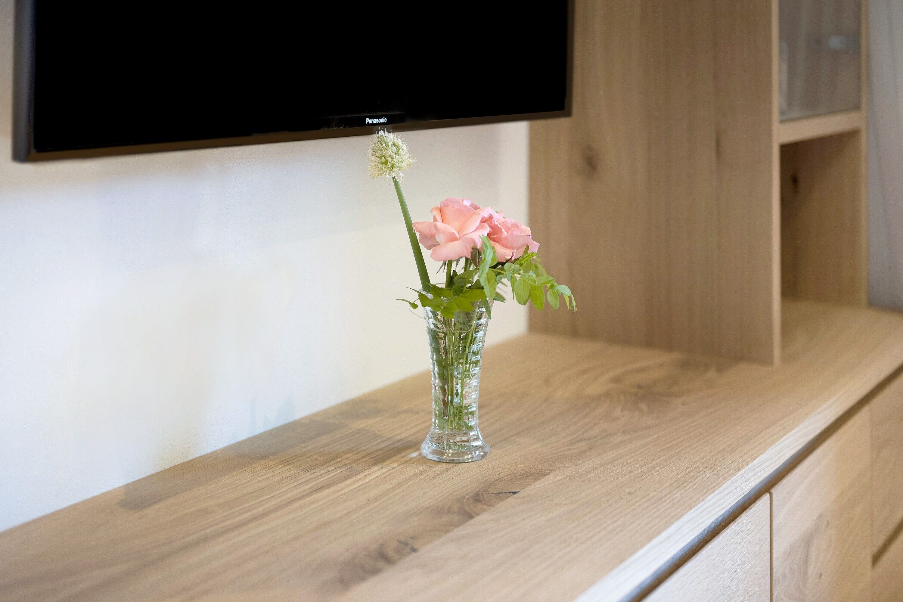 A wooden table with a clear glass vase containing a pink rose and a small white flower.