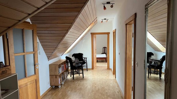 A narrow hallway with wooden flooring and a sloped ceiling. There are two chairs and a small table in the center, with a bed visible in the background.