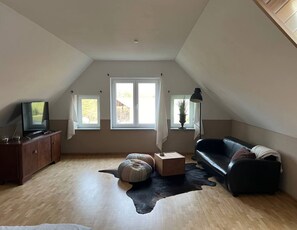 A spacious attic room with a sloped ceiling, a wooden floor, a black leather sofa, a small wooden coffee table, a television on a wooden cabinet, and two windows with white curtains.