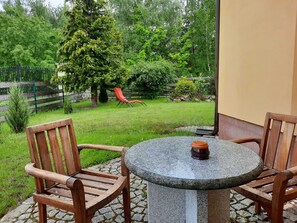 A patio with a round stone table and wooden chairs, overlooking a garden with a red chair and various plants.