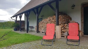 A covered outdoor seating area with two red chairs, a table, and a bench. A stack of firewood is visible in the background.