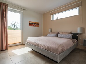 A bedroom with a large bed, a window with blinds, a painting on the wall, and a nightstand with a lamp.