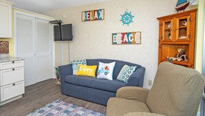 A living room with a blue sofa, a tan armchair, a wooden cabinet, and wall decorations including 'RELAX' and 'BEACH' signs.