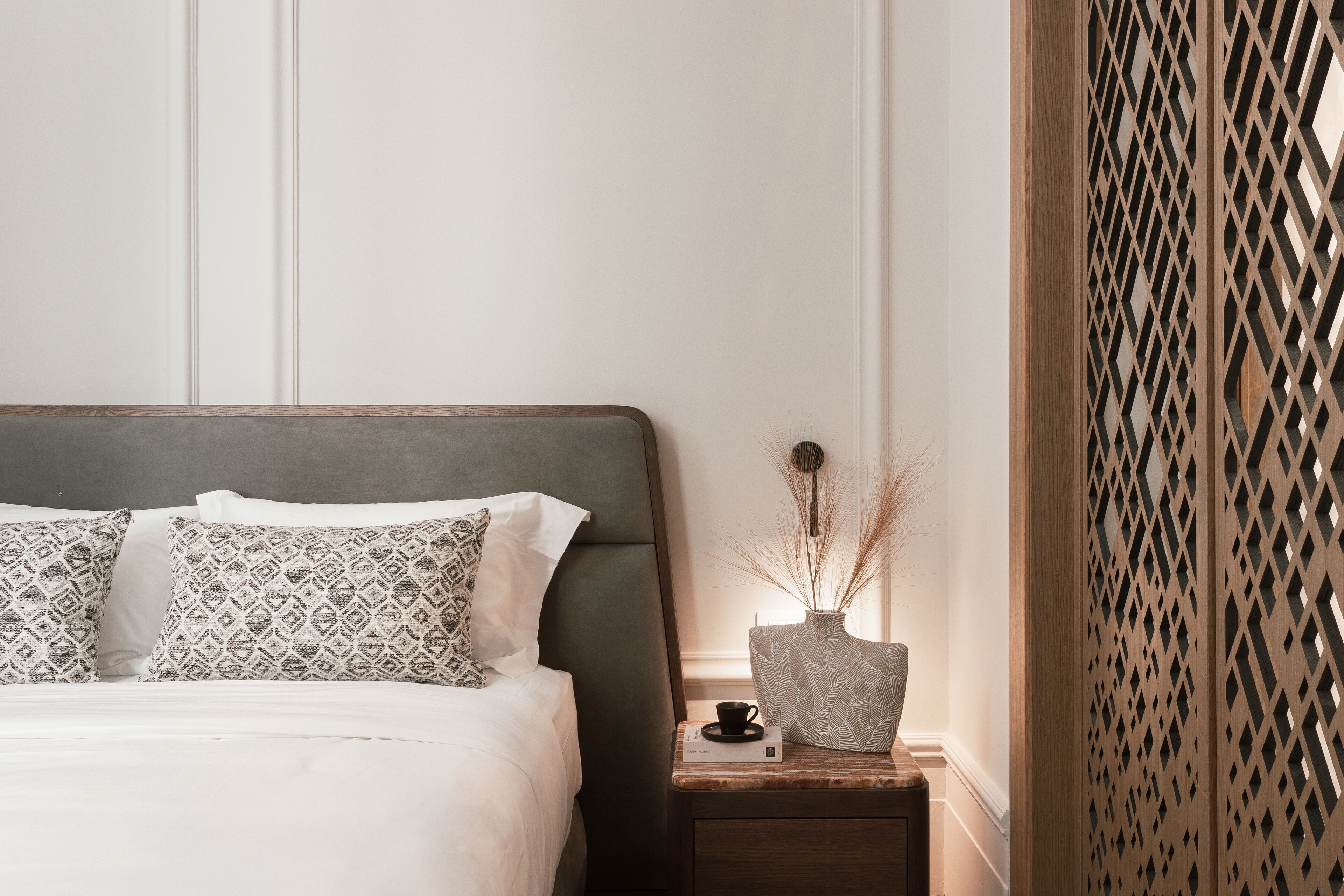 Una cama bien hecha con almohadones estampados, una mesita de noche con un florero y una taza, y un panel de madera decorativo.