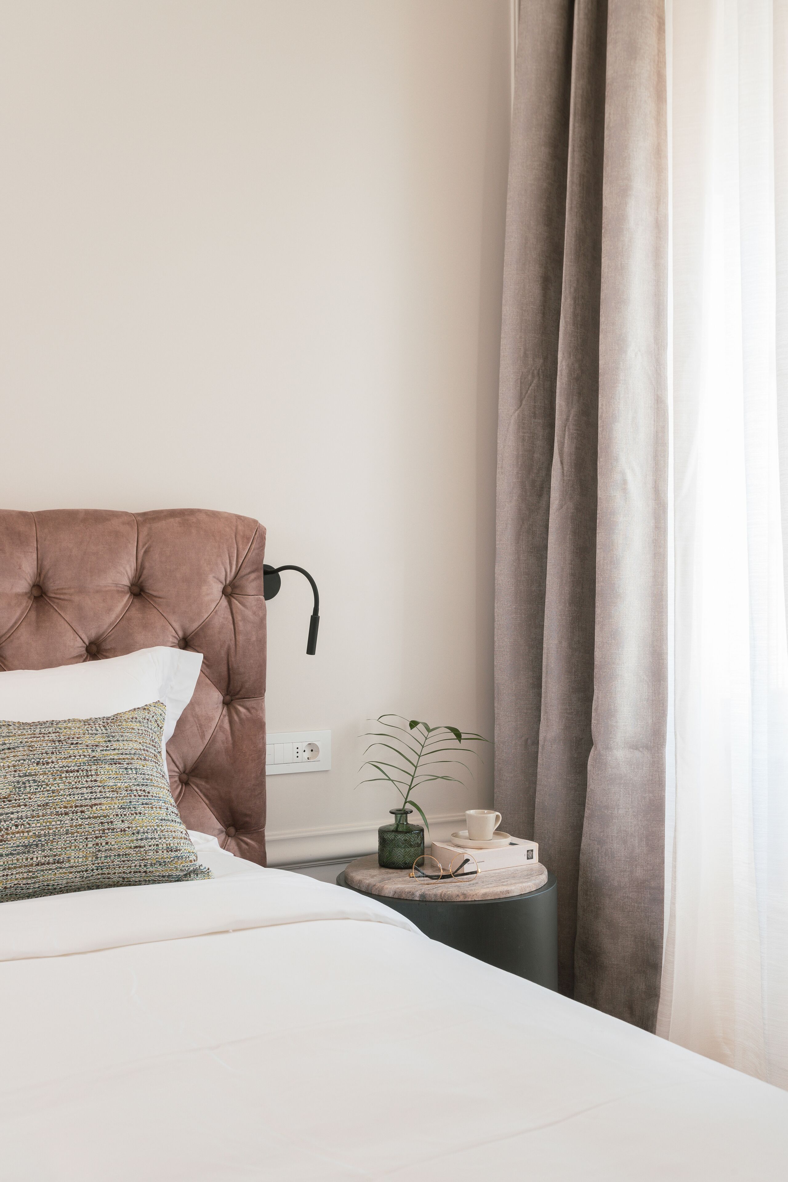 A neatly made bed with a tufted headboard, a bedside table with a cup and a small plant, and a window with curtains.