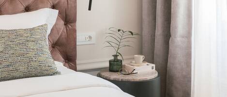 A neatly made bed with a tufted headboard, a bedside table with a cup and a small plant, and a window with curtains.