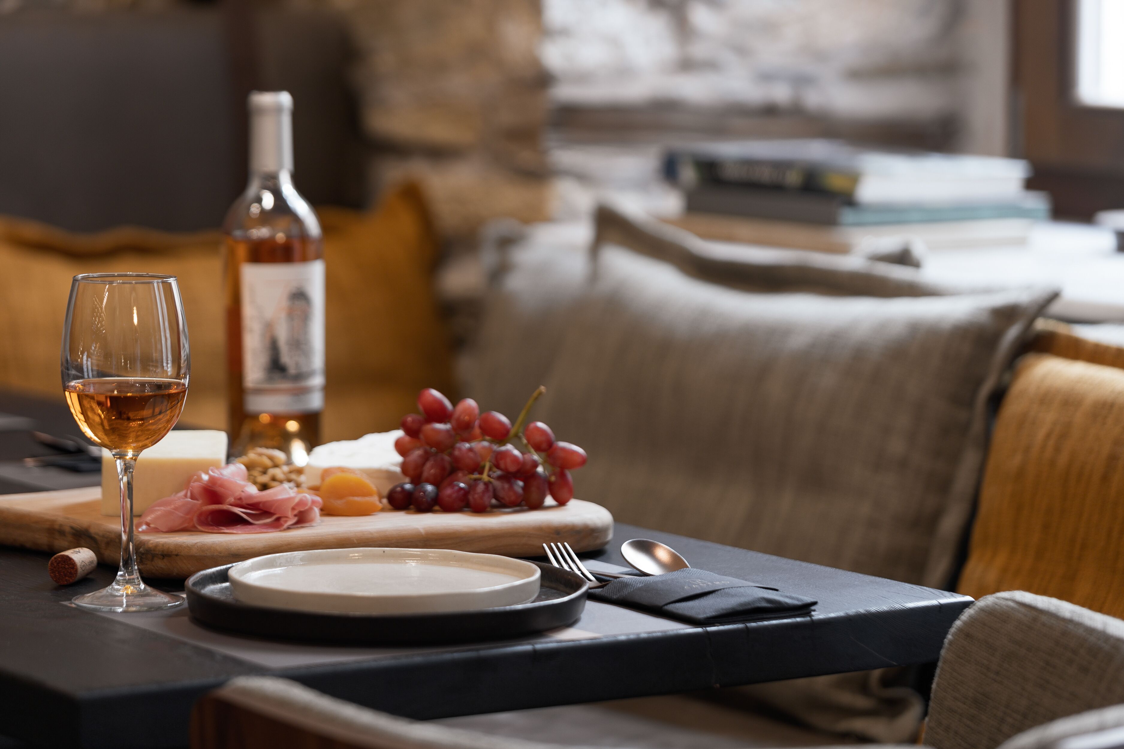 Una mesa con vajilla, uvas, queso y una copa de vino junto a una botella.