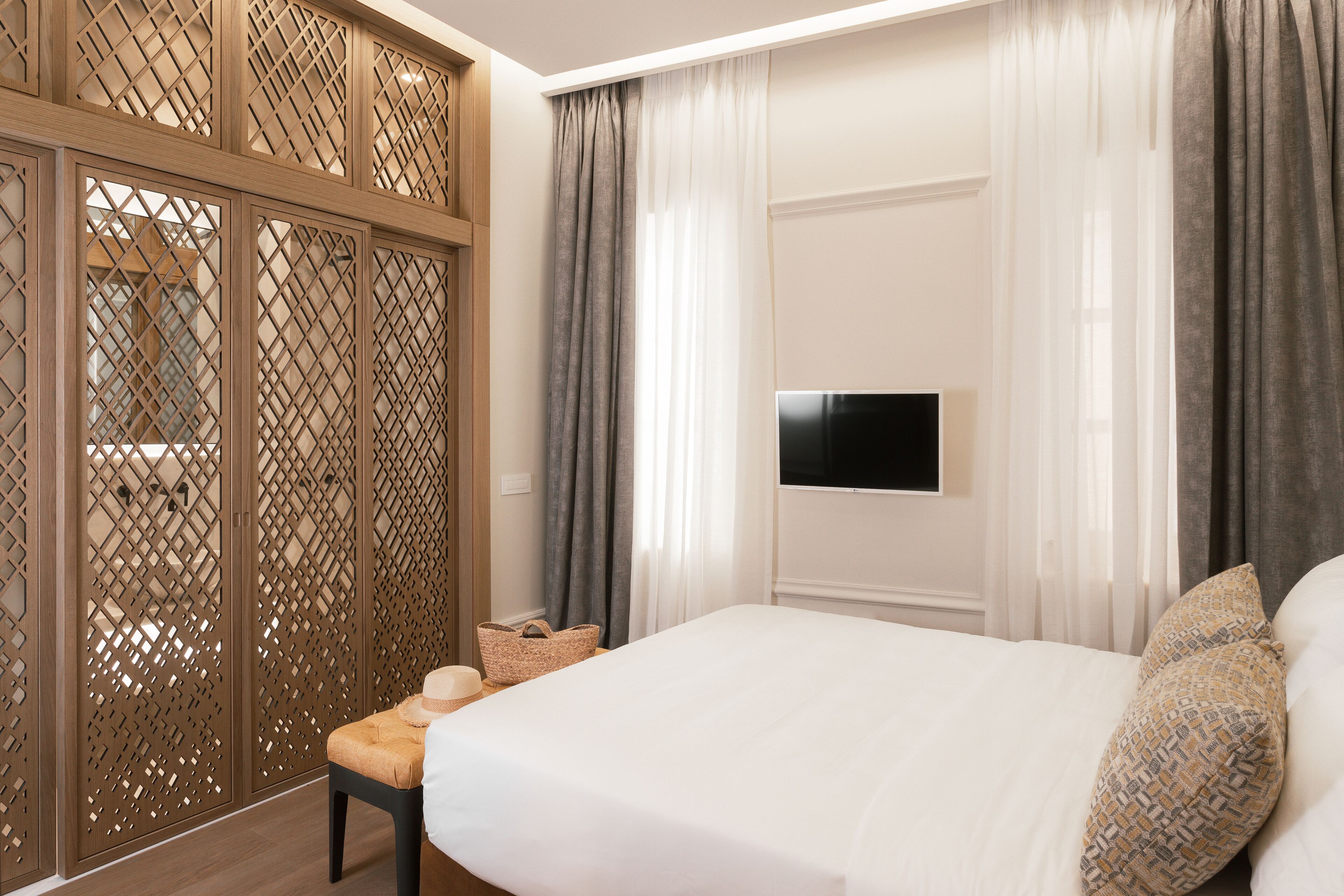 Una habitación de hotel con una cama, un televisor montado en la pared, un separador de madera y un banco con una cesta.